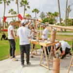 Team of people in white shirts assembling a large, homemade launch device outdoors, with palm trees in the background.