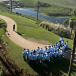 Bird's-eye view of a group of people in blue shirts standing in a semicircle on a curved park path near a pond, with a person and a small drone on the ground nearby.