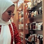 Woman in a white hijab and red polka-dot dress browsing perfume bottles on a store shelf in a cosmetics shop