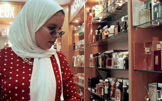 Woman in a white hijab and red polka-dot dress browsing perfume bottles on a store shelf in a cosmetics shop