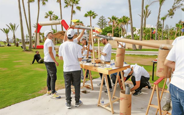 Team of people in white shirts assembling a large, homemade launch device outdoors, with palm trees in the background.